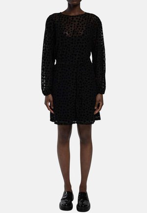 Femme portant une robe noire à manches longues avec des motifs de cœurs en tissu transparent, une ceinture à la taille et des mocassins noirs brillants, debout devant un fond uni.