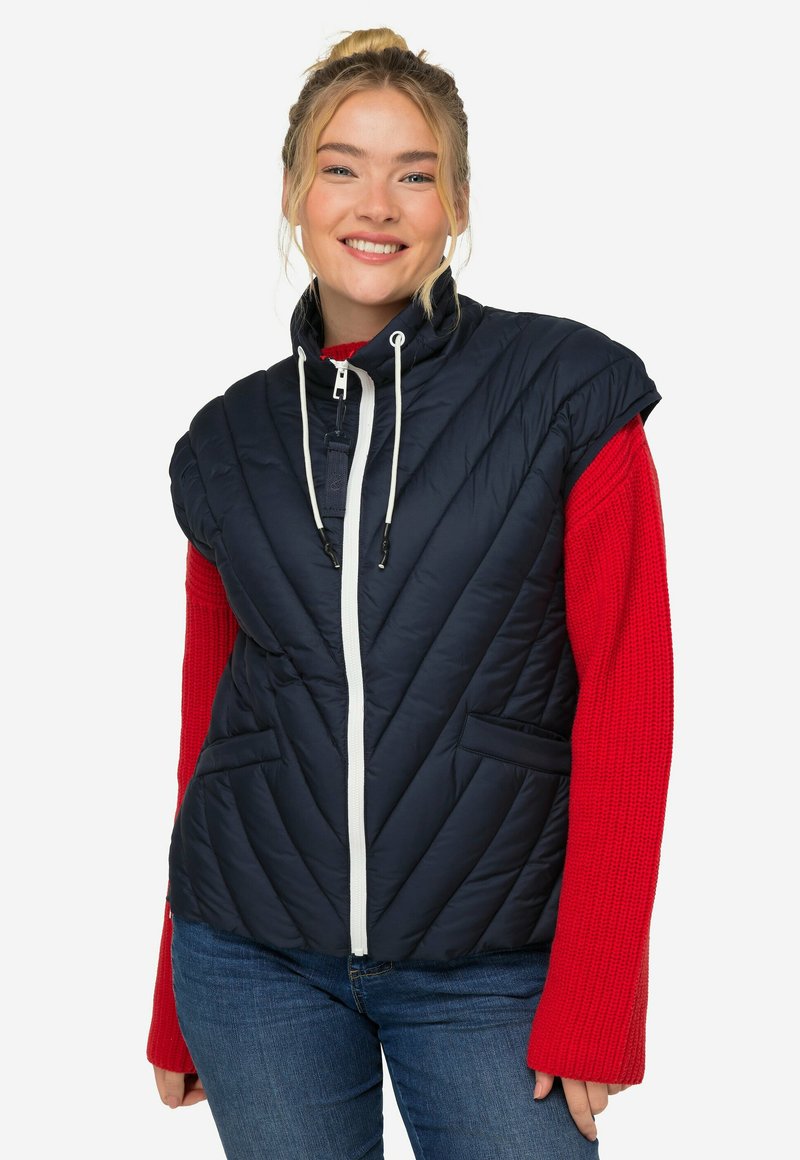 Marinblå quiltad väst med hög krage, vit dragkedja och två framfickor, kombinerad med en röd ribbad långärmad tröja.