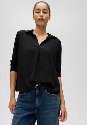 Frau trägt eine schwarze langärmelige Bluse mit V-Ausschnitt und blaue Jeans, steht vor einem einfachen hellen Hintergrund.