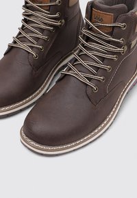 Botas de cuero marrón hasta el tobillo con detalles en marrón claro, cuello acolchado y cordones beige. Presentan un acabado texturizado y costuras blancas en contraste en la suela.