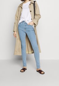 Beige trenchcoat med knappar, vit t-shirt, ljusblå skinny jeans och vinröda sandaler. Släta tygtexturer och smal passform.