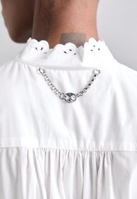 Biela bavlnená blúzka s vlnkovaným golierom a výstrihom so záhybmi na chrbte. Na krku je pripevnený strieborný reťazec s kruhovým detailom.