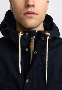Navy blue jacket with a hood, featuring leather accents, metal snaps, and a front zipper. Thick white drawstrings add detail.