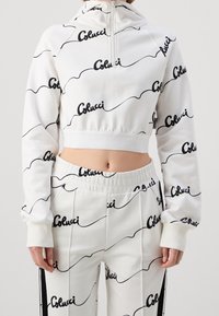Haut de hoodie blanc court avec un pantalon assorti, portant une impression du logo noir "Colucci", montrant le ventre et avec des manches de coupe décontractée.