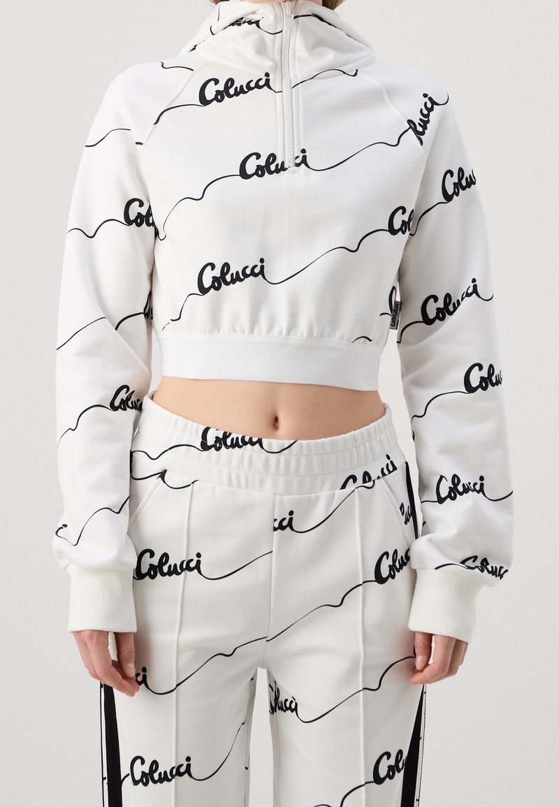 Haut de hoodie blanc court avec un pantalon assorti, portant une impression du logo noir "Colucci", montrant le ventre et avec des manches de coupe décontractée.