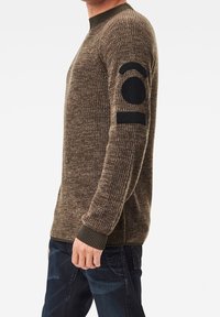 Mann trägt einen braunen Strickpullover mit schwarzem Logo-Patch auf dem oberen Ärmel, kombiniert mit dunkelblauen Jeans, steht seitlich vor einem weißen Hintergrund.