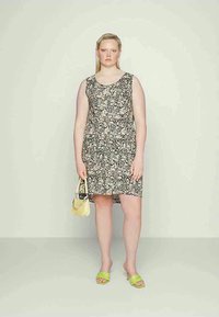 Mulher vestindo um vestido sem mangas preto e branco com estampado floral, segurando uma pequena bolsa amarela e usando sandálias de salto verde-lima.