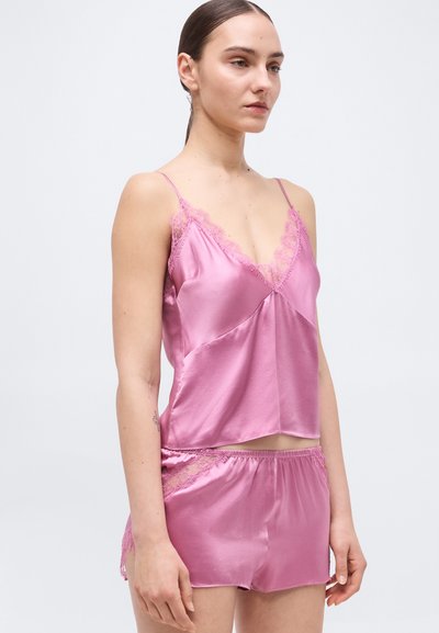 Γυναίκα που φοράει ροζ σατέν καμιζόλ και σορτς που ταιριάζουν με δαντελένια διακόσμηση, στέκεται μπροστά σε απλό φωτεινό φόντο, κοιτώντας ελαφρώς προς τα δεξιά.
