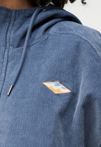 Κοντινό πλάνο ενός μπλε καπαρντινέ φούτερ με κουκούλα με ένα μπαλώμα σε σχήμα ρόμβου που φέρει λευκά, μπλε και κίτρινα αφηρημένα κύματα στο στήθος.