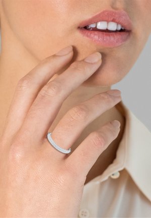 Mano con anello d'argento incastonato di diamanti che tocca il viso vicino alle labbra, indossa una camicia beige con colletto su sfondo neutro.
