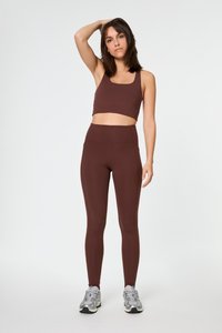 Smeđi sportski crop top i tajice visokog struka od rastezljivog materijala, upareni sa sivim trkačkim tenisicama. Glatka tekstura, minimalni šavovi.