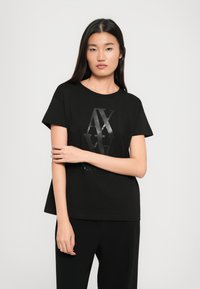 Aasia naine, kellel on pikad tumedad juuksed, kannab musta t-särki, millel on kergelt tõstetud läikiv "AX" logo, seistes ühtlasel heledal taustal.