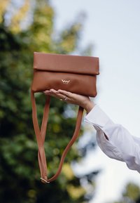 Hnědá kožená kabelka se sklápěcím vrškem, vybavená logem, nastavitelnými popruhy a hladkým povrchem, držena v ruce proti rozostřenému zelenému pozadí.