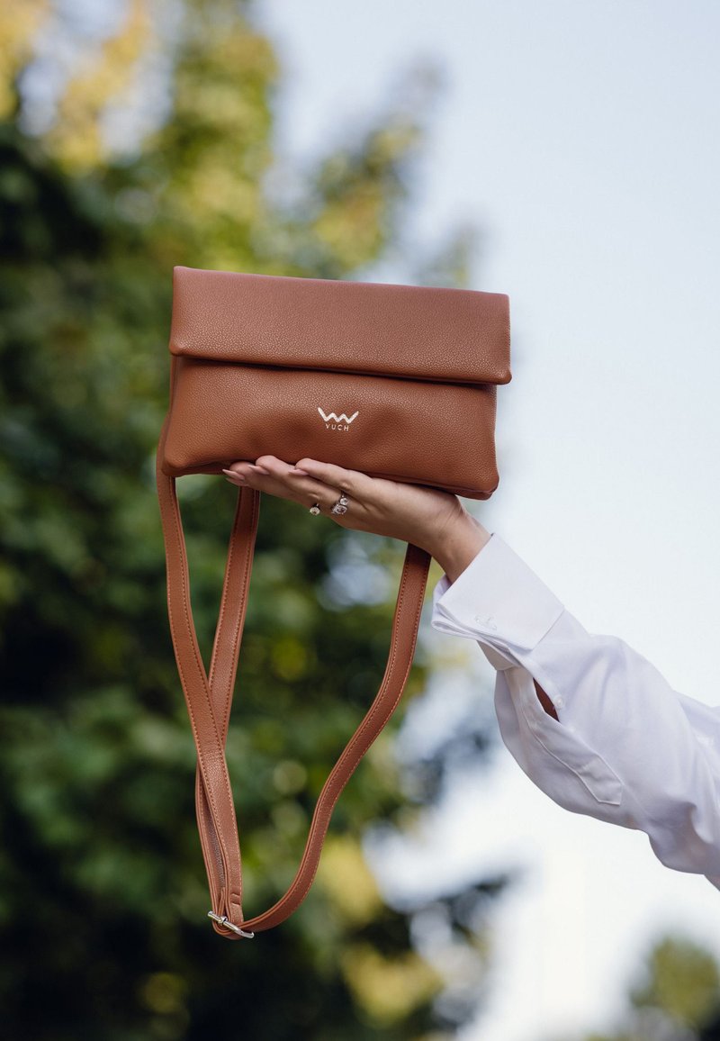 Hnědá kožená kabelka se sklápěcím vrškem, vybavená logem, nastavitelnými popruhy a hladkým povrchem, držena v ruce proti rozostřenému zelenému pozadí.