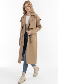 Jasno beżowy trench coat z podwójnym zapięciem, szerokim kołnierzem i klapami na ramionach, w połączeniu z jasnymi sznurowanymi botkami i niebieskimi dżinsami.