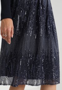 Robe marine en sequins avec des rayures verticales de sequins scintillants, présentant une silhouette fluide et une couche inférieure transparente.