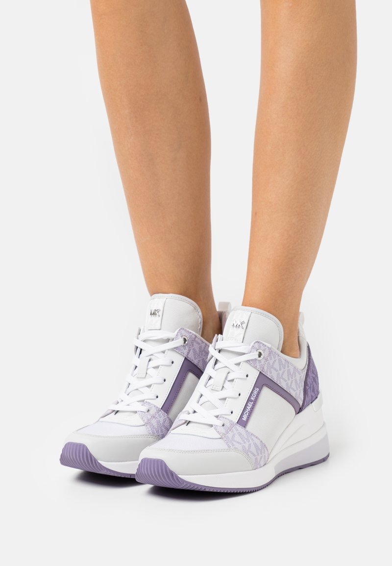 Gamabe con sneakers con zeppa Michael Kors bianche e lavanda con dettagli del marchio, lacci annodati, in piedi su una superficie bianca.