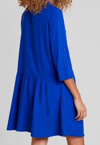 Robe bleue longueur genou avec une jupe froncée, des manches trois-quarts et des détails de boutons sur les manches. Fabriquée en tissu lisse.