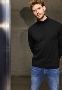 Svart stickad tröja med hög krage, kombinerad med ljusblå denimjeans. Tyget verkar mjukt, med en slät struktur och figurnära design.