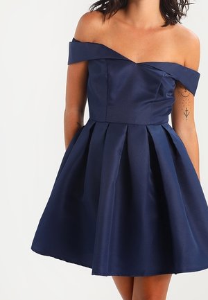 Marineblauwe off-shoulder jurk met geplooide rok, gedragen door een persoon met zichtbare tatoeages op de bovenarmen.