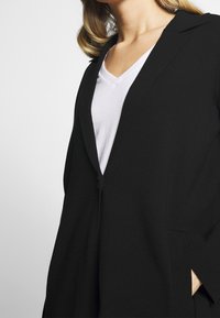 Svart blazer med skalkrage, enkel knappstängning och sidofickor. Bärs över en vit t-shirt med V-hals; slät textur.