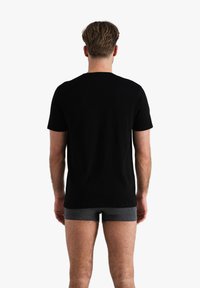 T-shirt en coton noir à manches courtes, vu de dos, porté sur un short de boxe gris. Design minimal, sans motifs ni logos visibles.