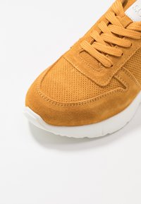 Brown suede sneaker with textured surface, featuring white rubber sole and matching laces, angled view highlighting the toe and upper detail.
