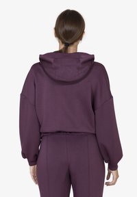 Felpa corta viola con cappuccio, dotata di spalle cadenti e orlo elasticizzato. Abbinata a pantaloni a vita alta coordinati.
