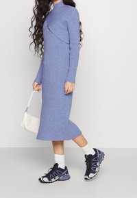 Vestido midi de canalé azul claro con un cuello alto y un frente con capas, combinado con zapatillas oscuras y un bolso blanco pequeño.