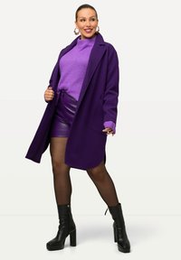 Lila ullkappa, ribbad lila tröja, glänsande lila shorts och svarta platåstövlar. Har stora örhängen och en slät textur.