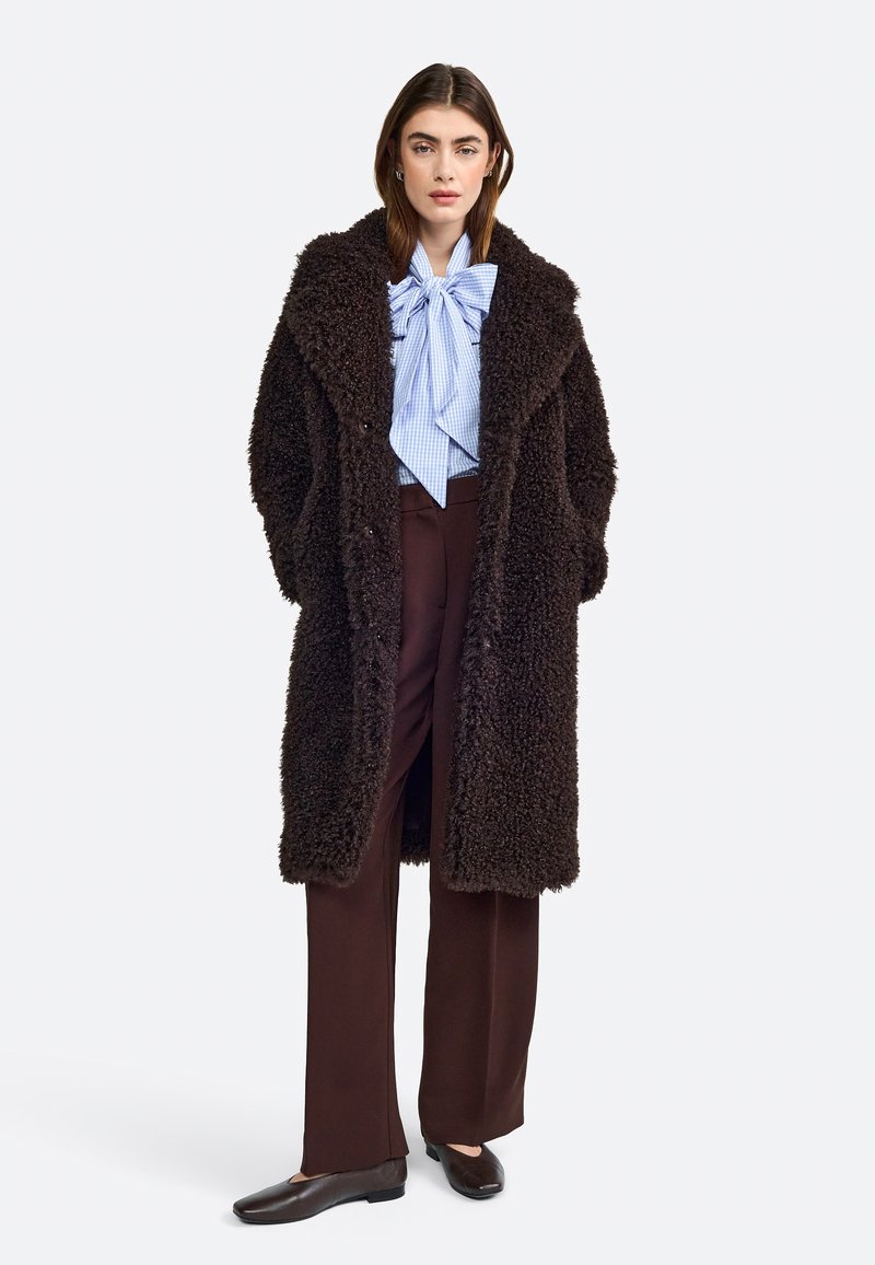 Cappotto lungo marrone e peloso con colletto, indossato sopra una blusa a righe blu con un grande fiocco. Abbinato a pantaloni marroni e scarpe scure.
