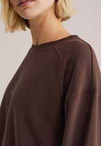 Ruda sportinė palaidinė su apvalia iškirpte ir raglan rankovėmis, pasižyminti minkšta tekstūra ir minimalistiniu dizainu, rodoma iš šono.