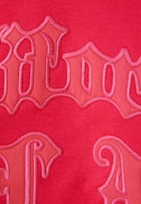 Červená látka s vyvýšeným, tonálním vyšíváním stylizovaného textu. Textura je hladká, s různými odstíny růžové pro kontrast.