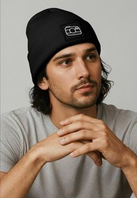 Young man with dark hair and beard wearing a black beanie with a white camera icon, resting hands and looking slightly to the side.