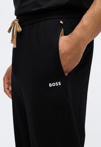 Crne atletske hlače s pojasom na vezivanje, bež detaljima, bočnim džepovima i bijelim "BOSS" logotipom na lijevom bedro. Glatka tekstura tkanine.