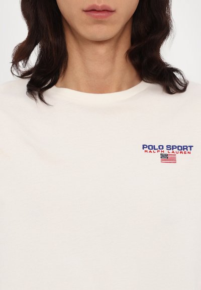 Kratka kremasta majica s kratkimi rokavi iz bombaža, s pisanjem "Polo Sport" v modri in rdeči barvi ter oblikovanjem ameriške zastave. Preprost kroj s krožnim izrezom.