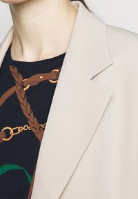 Beige kavaj med slag, visad över en svart skjorta med ett brunt kedjemönster med gulddetaljer och gröna inslag.