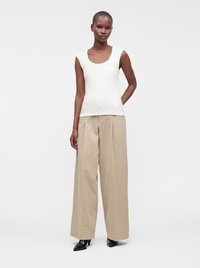 Femme portant un haut blanc sans manches, un pantalon beige à jambes larges et des chaussures noires à bouts pointus, debout devant un fond blanc uni.
