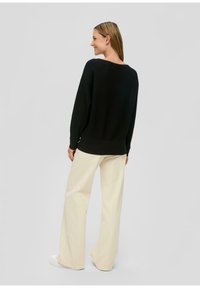 Schwarzer gerippter Pullover mit langen Ärmeln und weitem Ausschnitt, kombiniert mit locker sitzenden cremefarbenen Hosen und weißen Turnschuhen.