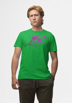 Grøn bomulds T-shirt med et struktureret lilla grafisk design. Korte ærmer, rund hals, afslappet pasform. Model poserer med hænderne i lommerne.