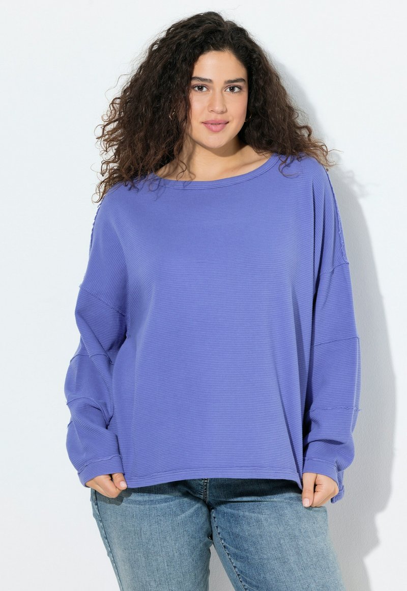 Mujer joven con cabello rizado, vestida con un suéter morado holgado y jeans de color azul claro, de pie contra un fondo blanco liso.