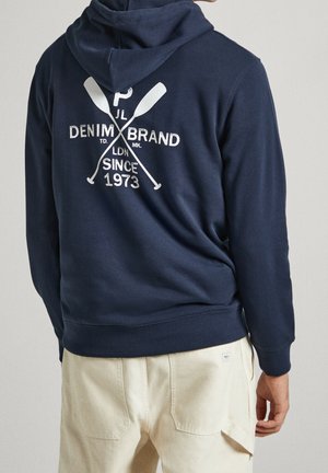 Persona con sudadera con capucha azul marino con remos cruzados y texto "Denim Brand Since 1973" en la espalda, combinada con pantalones beige claro.