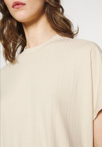 Beige ribbad topp med rund halsringning och korta ärmar, med en slät textur och subtila vertikala linjer.