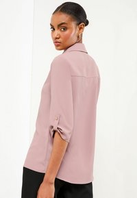 Blusa color rosa con maniche arrotolabili, dotata di un colletto classico e una singola cucitura posteriore. Tessuto liscio con minimalismo nei dettagli.