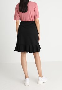 Gonna nera svasata con orlo arricciato, abbinata a una maglietta rosa a maniche corte. Il modello indossa delle sneakers bianche, mostrando un outfit casual di spalle.