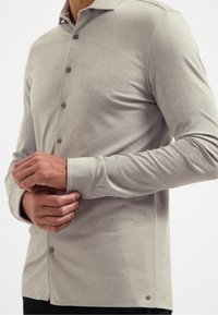 Lange mouwen shirt in lichtgrijs, gemaakt van zachte stof met een subtiele textuur. Bevat een puntige kraag en zeven knopen aan de voorkant.