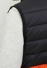 Czarna puchowa kamizelka z poziomym przeszyciem, bez rękawów, z kontrastową lamówką, noszona na jasnoszarej bluzie z okrągłym dekoltem.