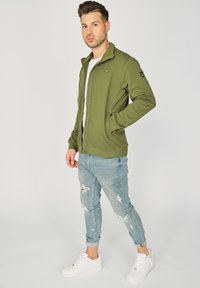 Oliwkowa kurtka zapinana na zamek z stójką, w połączeniu z jasnymi dżinsami z przetarciami i białymi trampkami.