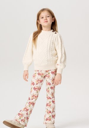 Pull en maille torsadée crémeux avec des manches bouffantes ; pantalon à motifs avec des designs floraux rouges et verts ; baskets beiges. Modèle se tenant avec assurance.