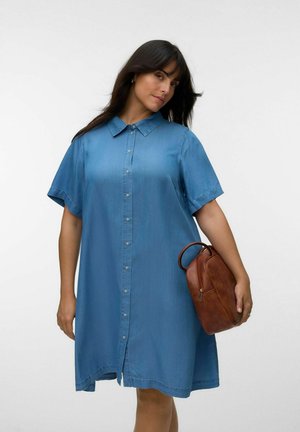 Femme portant une robe en jean bleue ample boutonnée, tenant un sac à main en cuir marron sur un fond uni.